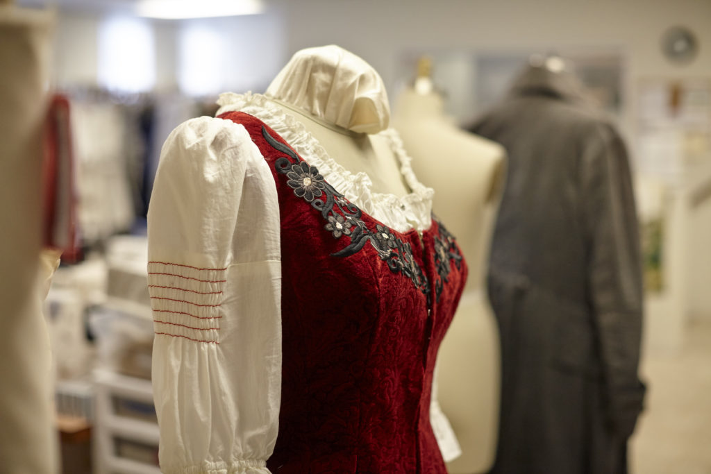 An intricately beaded red bodice is o verdressed on top of a puffy sleeved blouse on a bust, or judy, while empty busts sit in the background, waiting to be donned with costumes.